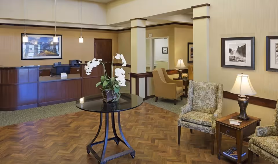 A senior living facility reception area with a wooden front desk, a round table with a white orchid plant in the center, patterned armchairs, side tables with lamps, framed pictures on the walls, and warm lighting.