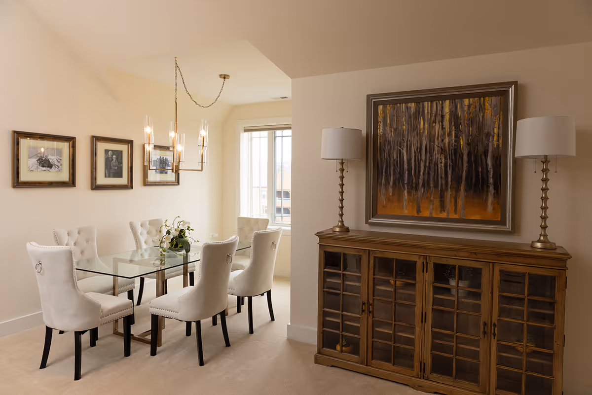 Bright dining room with a glass table and six upholstered chairs, a chandelier, framed photos on the wall, and a wooden sideboard topped with lamps and a large painting.