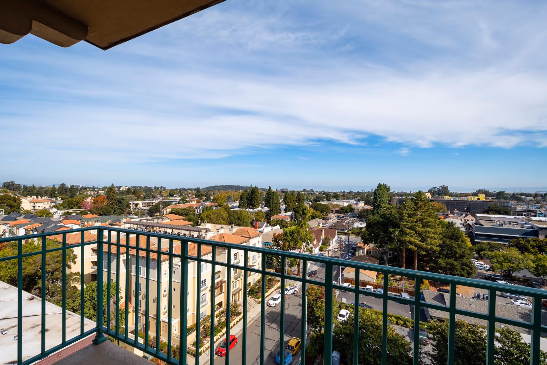 View from a balcony with a green railing overlooking a neighborhood of houses, trees, and a wide sky.