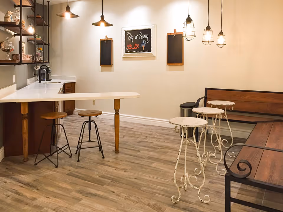 A cozy interior space featuring a small kitchen counter with two wooden stools, hanging pendant lights, and a seating area with a wooden bench and three small round tables with ornate metal legs. The wall has a framed chalkboard sign that reads 'Sip n' Scoop' and two smaller blank chalkboards on either side.
