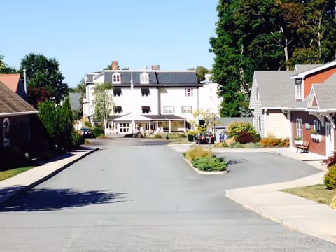 View of a paved road leading to a large white multi-story building with a porch, surrounded by smaller buildings and trees on a sunny day.