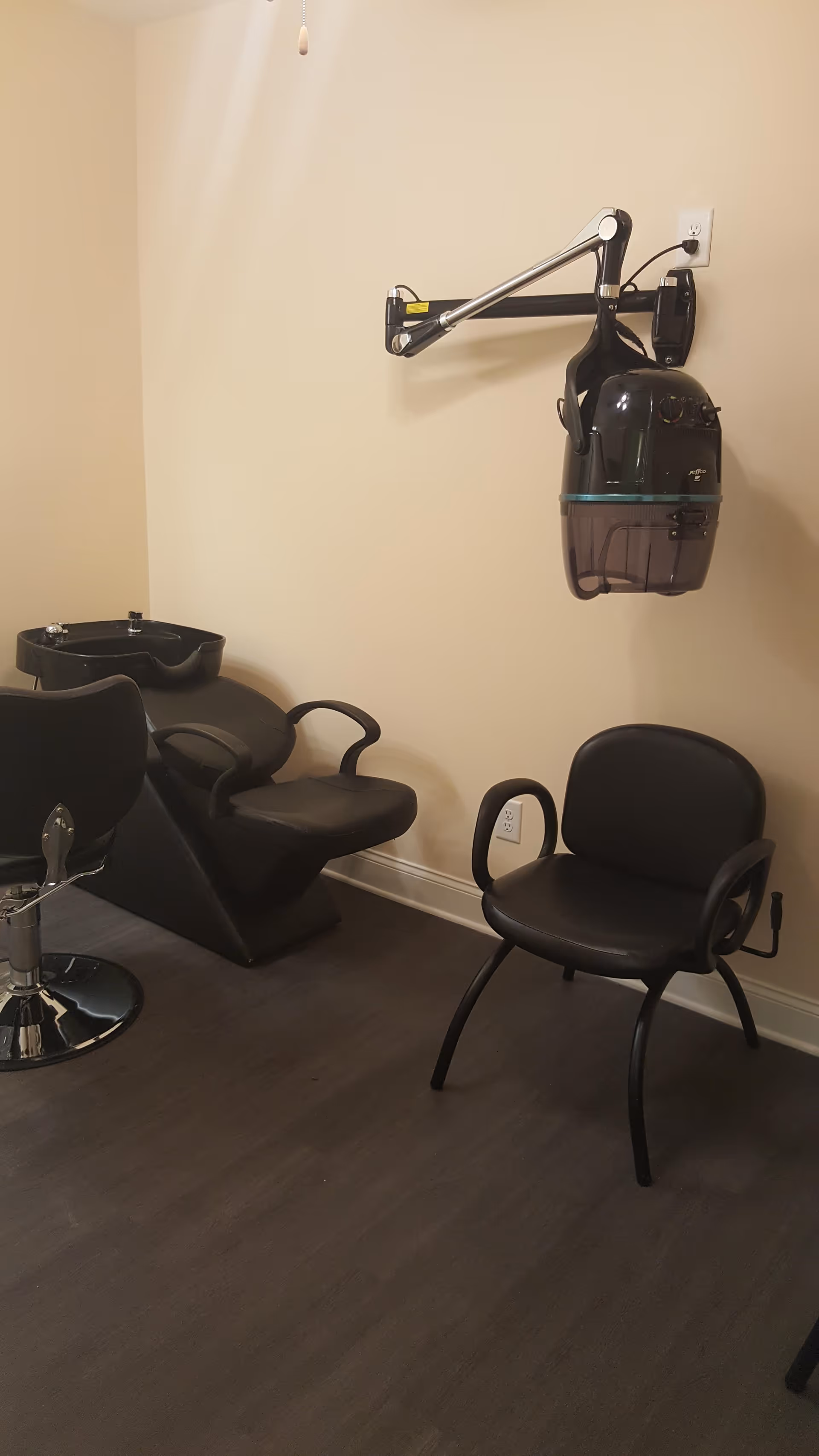 Interior view of a hair salon area with a black salon chair, a black shampoo station with a sink, a black waiting chair, and a wall-mounted hair dryer.