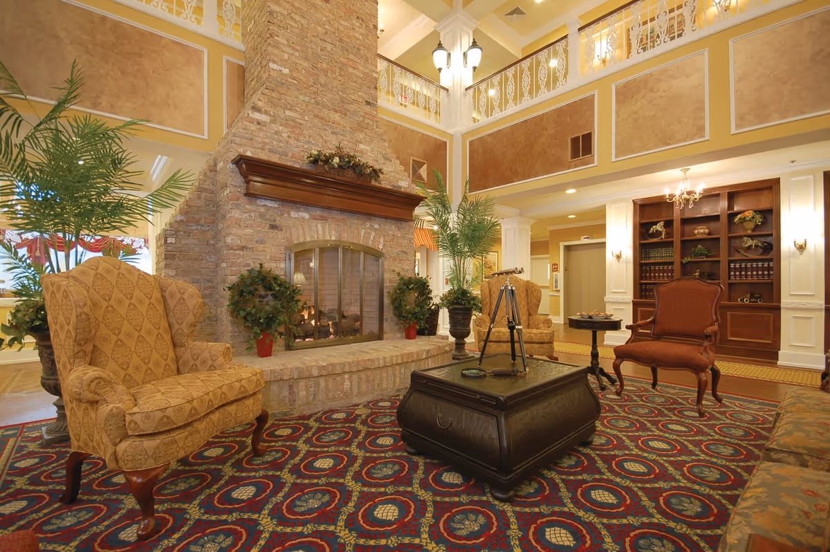 A spacious and elegant living room area with a large brick fireplace in the center. The room features high ceilings with decorative molding and a balcony railing above. There are several upholstered armchairs and a dark wooden coffee table with a decorative telescope on it. The floor is covered with a patterned carpet, and there are potted plants and a built-in wooden bookshelf with decorative items and books. Warm lighting enhances the cozy atmosphere.