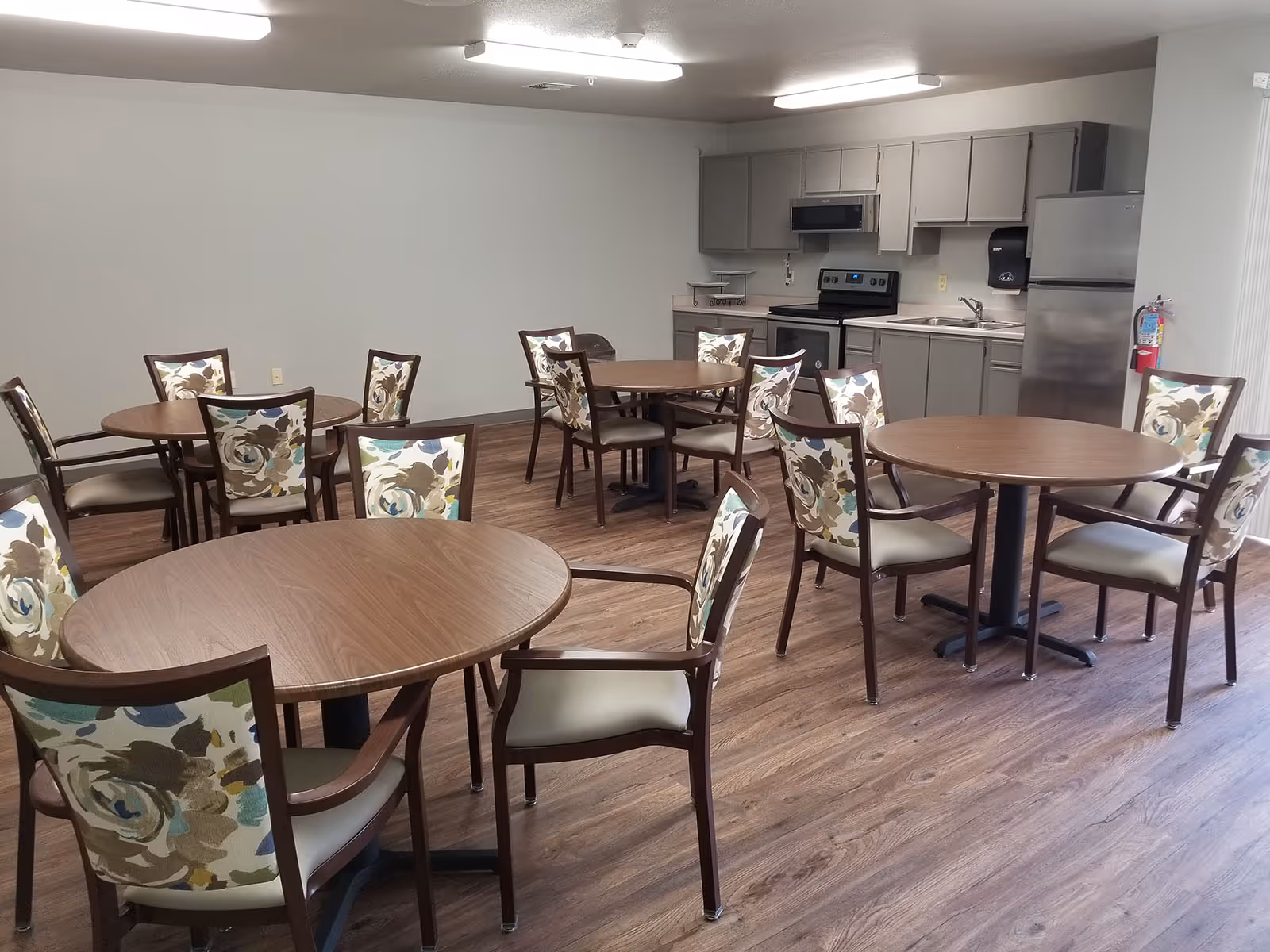 Communal dining room with round wooden tables, floral-upholstered chairs, and a kitchenette along the back wall.