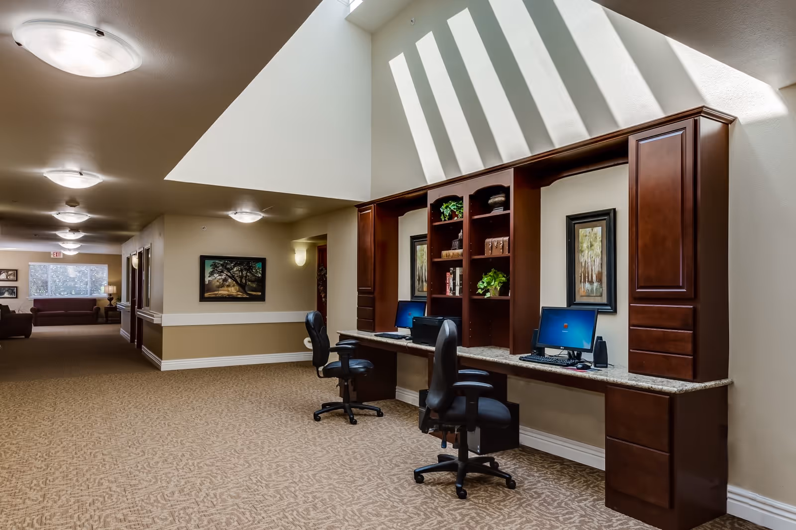 Interior hallway of Autumn View Gardens Assisted Living and Memory Care featuring a built-in wooden desk area with two computer workstations and office chairs. The space is well-lit with ceiling lights and natural light coming from a skylight above. The hallway has beige carpet and walls decorated with framed artwork, and there are seating areas visible in the background.