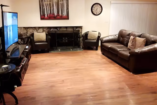 A living room with wooden flooring, a brown leather couch with two pillows, two black armchairs with light-colored cushions, a stone fireplace with a black screen, a wall clock, a painting of trees with red accents above the fireplace, and a TV on a stand to the left.