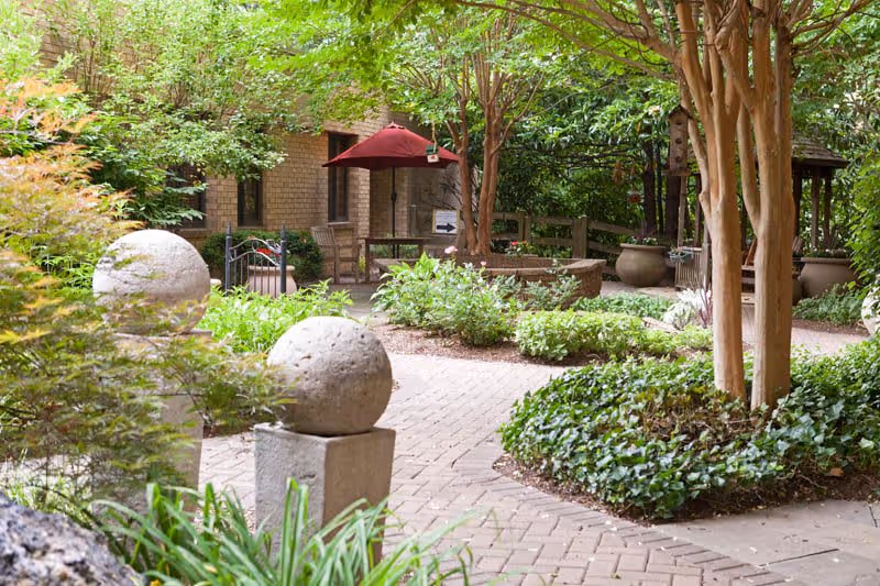 A peaceful outdoor garden area with a paved walkway winding through lush greenery, trees, and shrubs. There are two stone spheres on square pedestals in the foreground, a wooden bench with a red umbrella in the background, and a small gazebo partially visible on the right side.