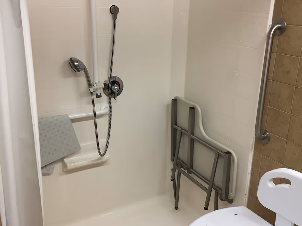 Accessible shower stall with a handheld showerhead, grab bars and a folded shower chair.