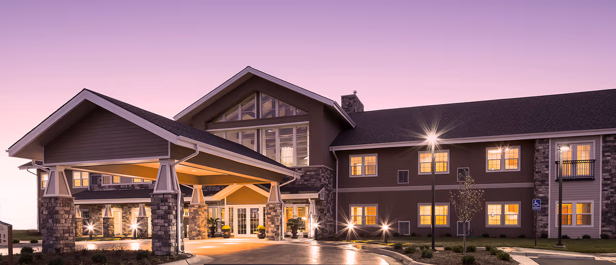 Exterior view of The Oxford Grand Assisted Living & Memory Care building at dusk with warm lights glowing from the windows and a covered entrance supported by stone pillars.