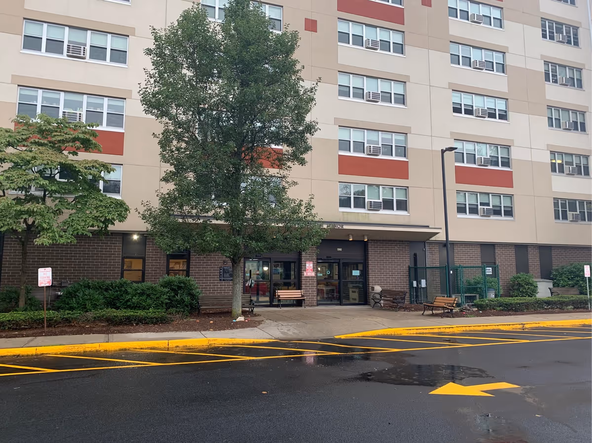 Exterior view of a multi-story building with beige and red accents, featuring multiple windows and air conditioning units. There is a sidewalk with benches, trees, and bushes in front of the entrance. The ground is wet, and there are yellow painted lines and an arrow on the asphalt road leading to the entrance.