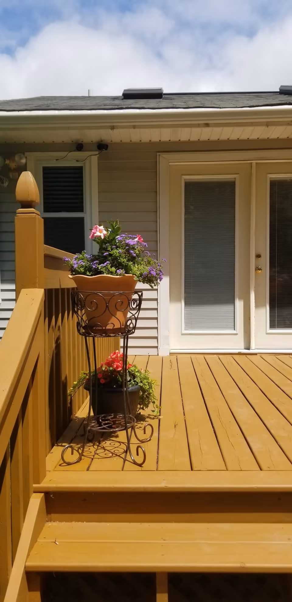 A wooden deck with two steps leading up to a set of white double doors with blinds. On the deck, there is a decorative metal plant stand holding a terracotta pot with purple and pink flowers, and a black pot with red flowers placed behind it. The exterior wall of the building is light gray with a small window to the left of the doors. The sky is partly cloudy.