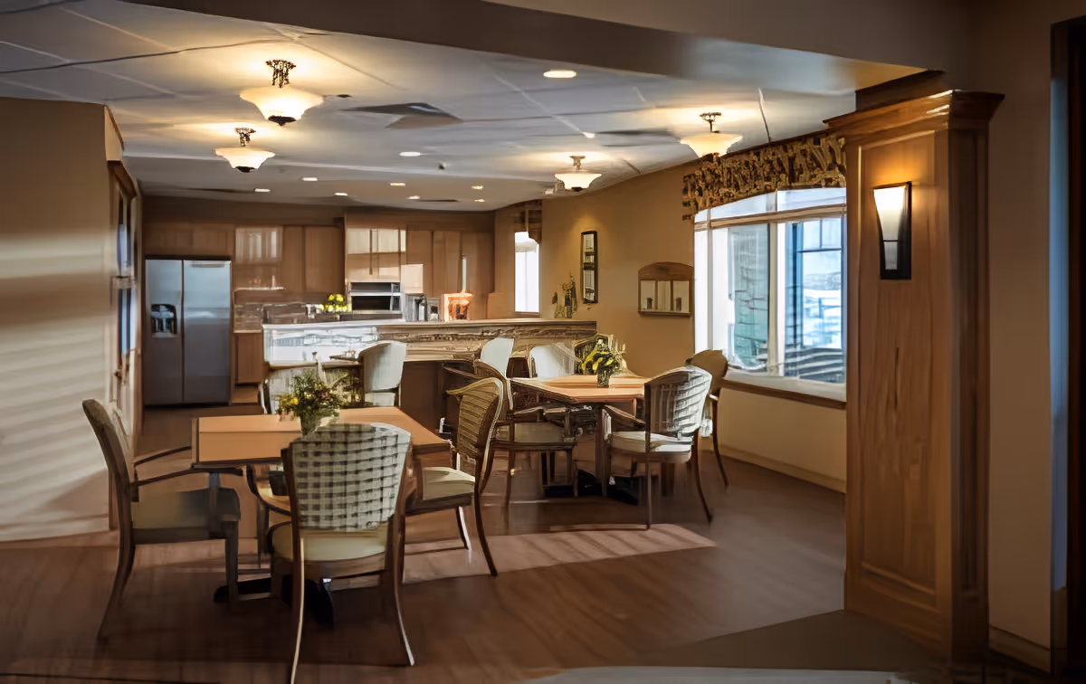 A cozy dining area in a retirement community featuring several wooden tables with cushioned chairs arranged around them. The room has warm lighting with ceiling fixtures and wall sconces. In the background, there is a kitchen area with wooden cabinets, a refrigerator, and a countertop bar with additional seating. Large windows on the right side allow natural light to enter the space.