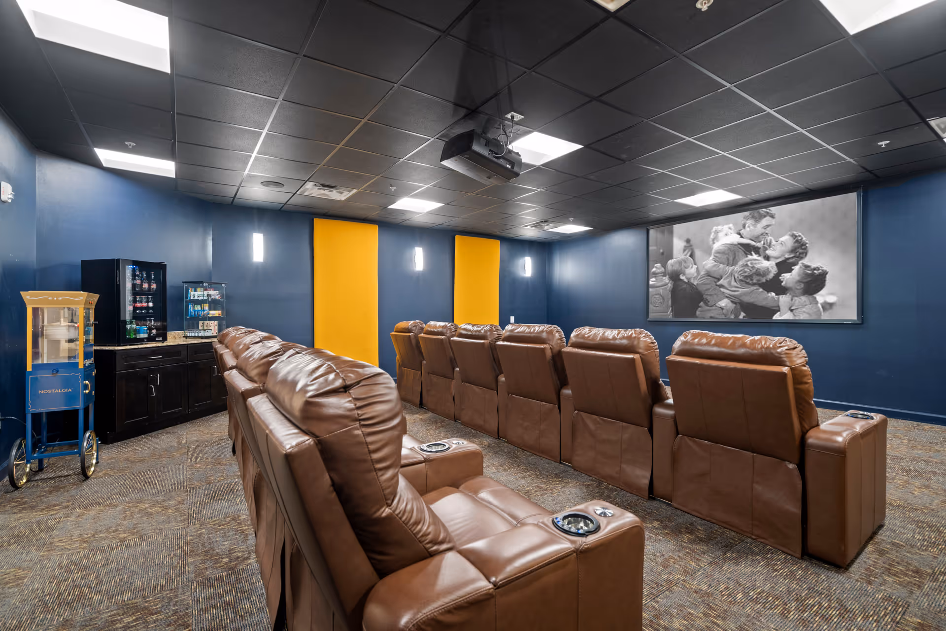 A cozy movie theater room with two rows of brown leather recliner chairs facing a large screen displaying a black and white movie scene. The walls are painted dark blue with yellow acoustic panels, and there is a popcorn machine and snack vending machines in the corner.