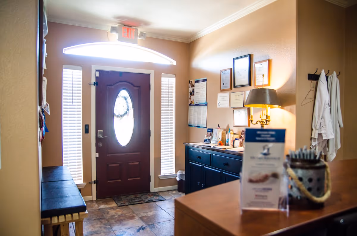Interior entry/reception area with a closed front door flanked by tall windows, a sideboard with framed certificates and a lamp, and a front desk with brochures.