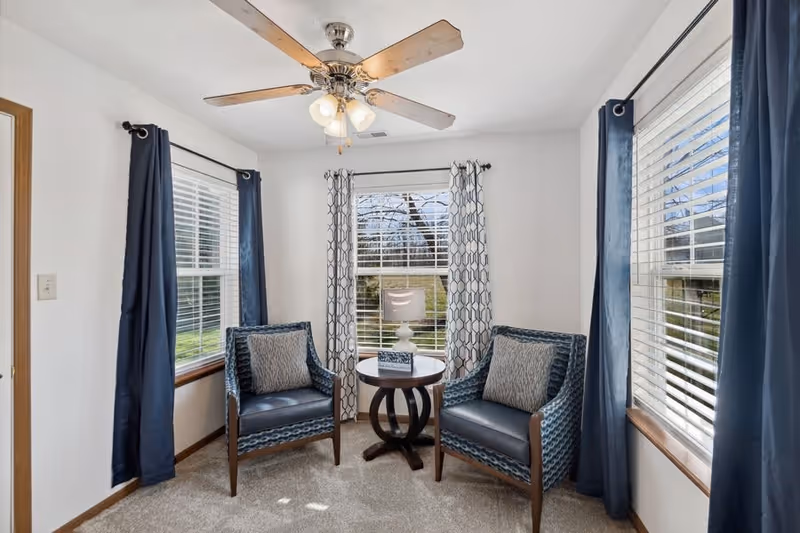 A small sitting area with two blue patterned armchairs and a round wooden table between them. The table holds a white lamp and a decorative box. The room has three windows with white blinds and blue curtains, and a ceiling fan with lights above. The walls are white and the floor is carpeted.