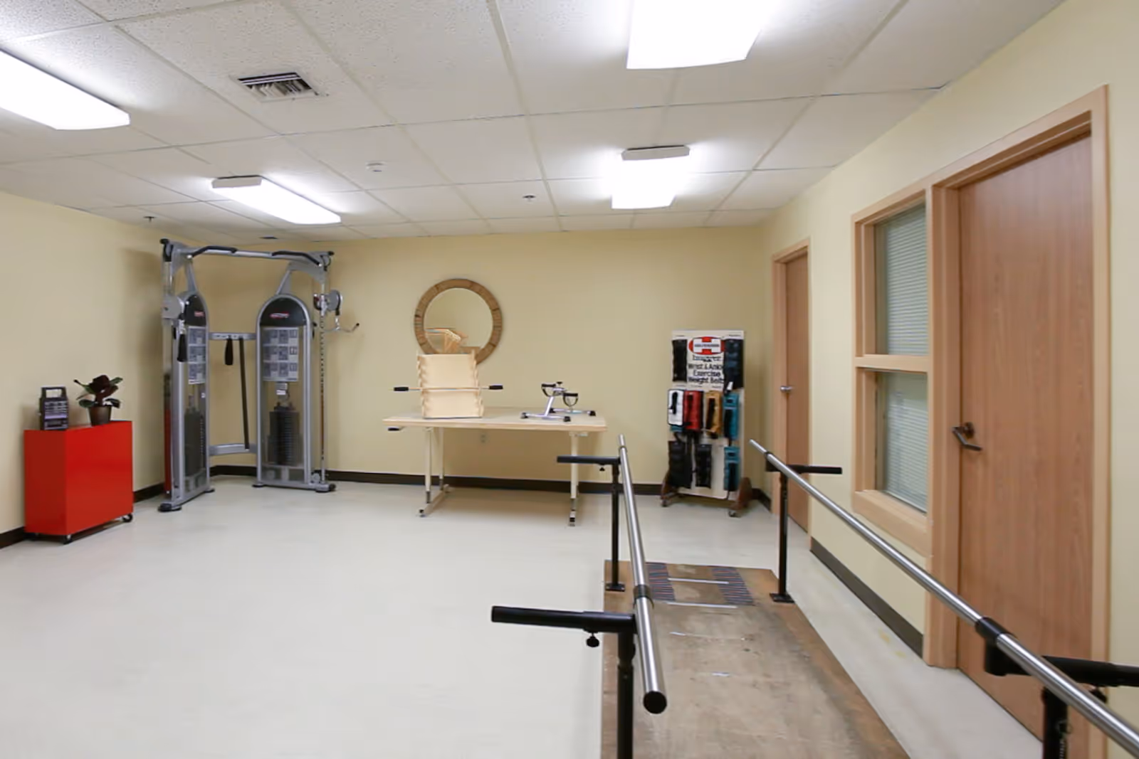 A rehabilitation or physical therapy room with parallel bars for walking exercises, a multi-functional exercise machine, a table with therapy equipment, a round wall mirror, and a display rack with medical supplies. The room has beige walls, a white ceiling with fluorescent lights, and two wooden doors with windows.