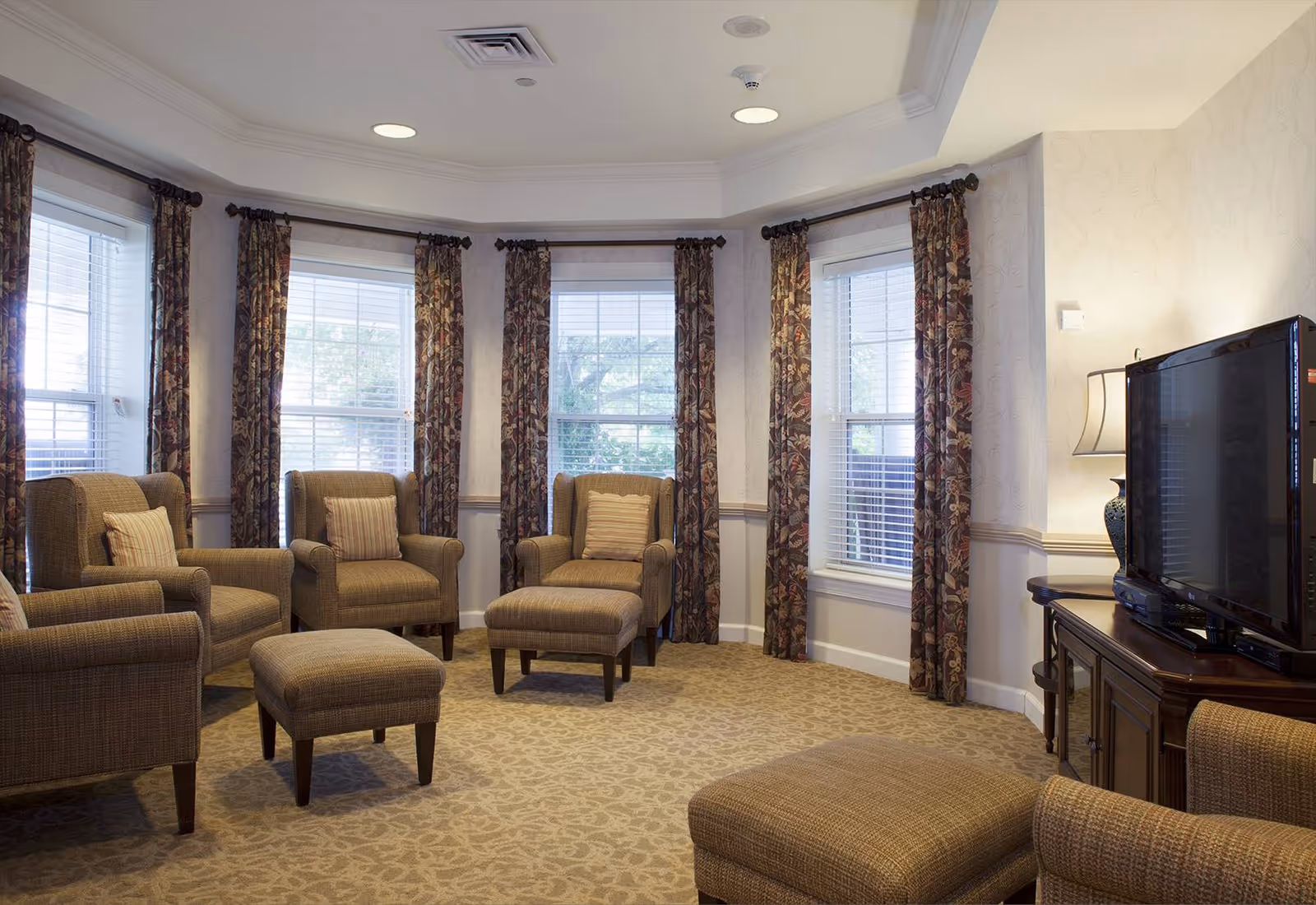 A cozy living room with multiple upholstered armchairs and matching ottomans arranged in a semi-circle. The room features large windows with floral patterned curtains, beige carpet with a subtle pattern, and a flat-screen TV on a wooden cabinet with a decorative lamp beside it.