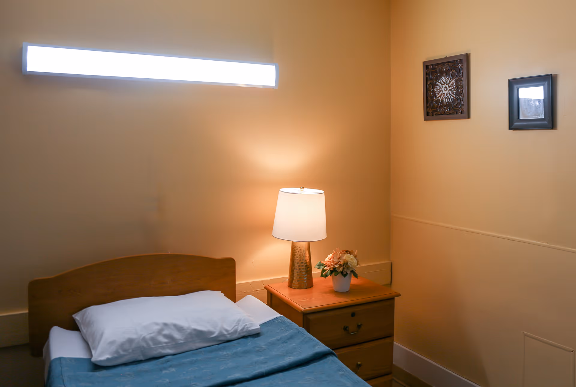 A simple bedroom with a single wooden bed covered with a blue blanket and a white pillow. Next to the bed is a wooden nightstand with a lamp and a small flower pot. The walls are painted beige and have two framed decorative pieces hanging on them.