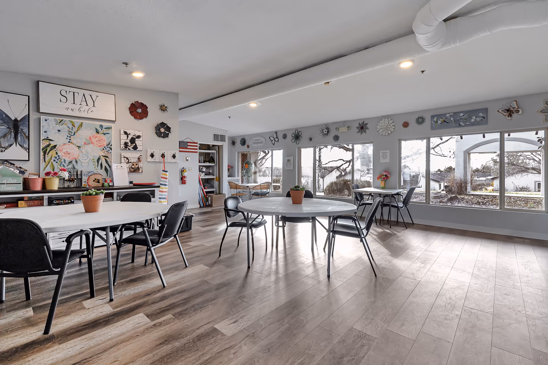 A bright and spacious common area in a senior living facility with round and rectangular tables surrounded by black chairs. The room features large windows letting in natural light, decorative wall art including flowers, butterflies, and inspirational signs, and a wooden floor. There are potted plants on the tables and shelves with books and decor items along the walls.