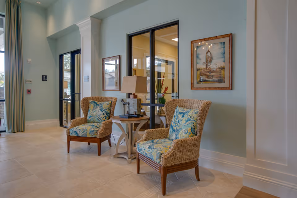 Two wicker armchairs with patterned cushions flanking a small table and lamp in a bright reception/lounge area.