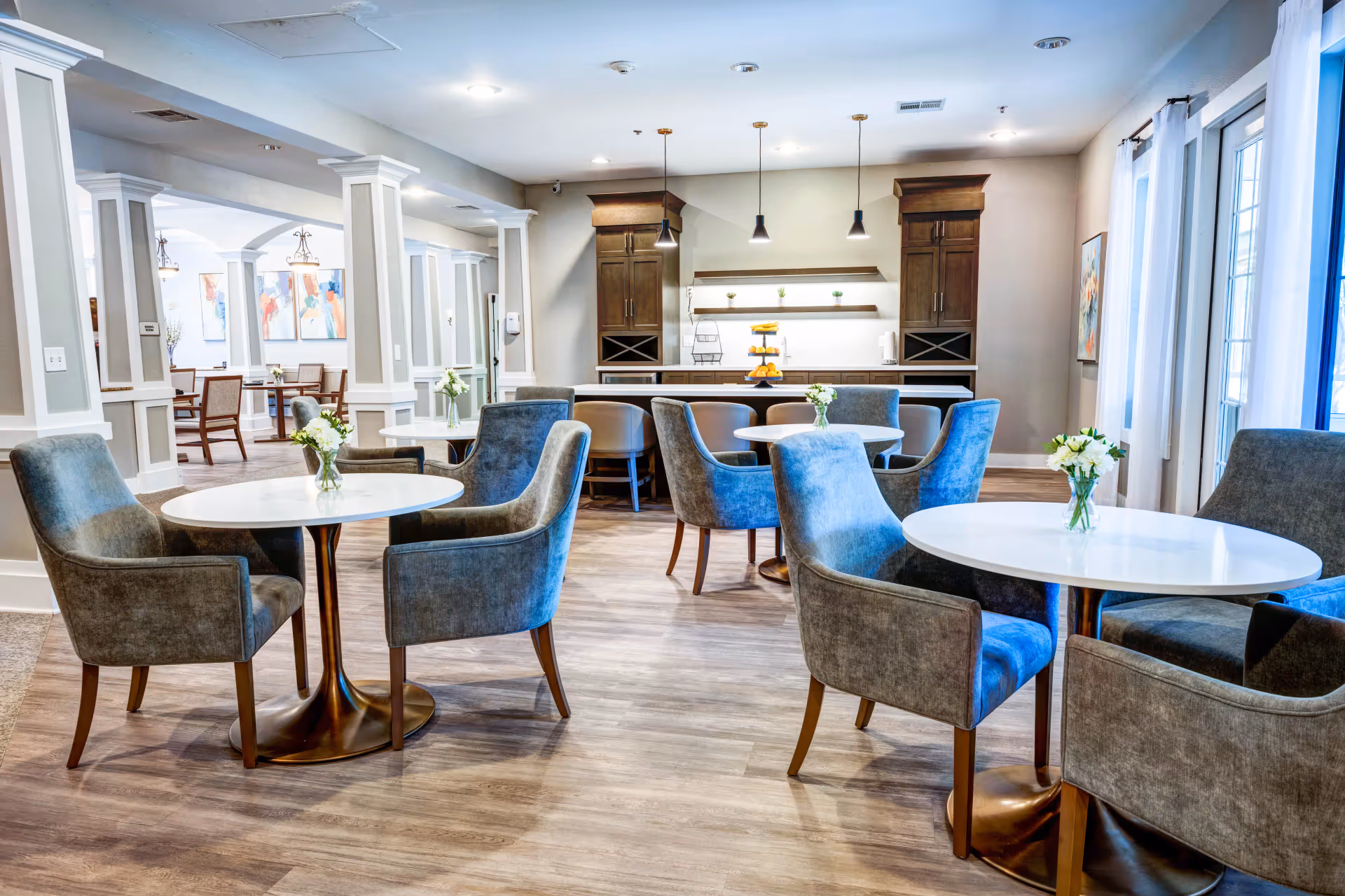 A bright and modern dining area in an assisted living facility featuring round white tables with gray upholstered chairs. Small flower vases are placed on each table. The room has wood flooring, pendant lights hanging over a counter with dark wood cabinets, and large windows with white curtains allowing natural light to enter.