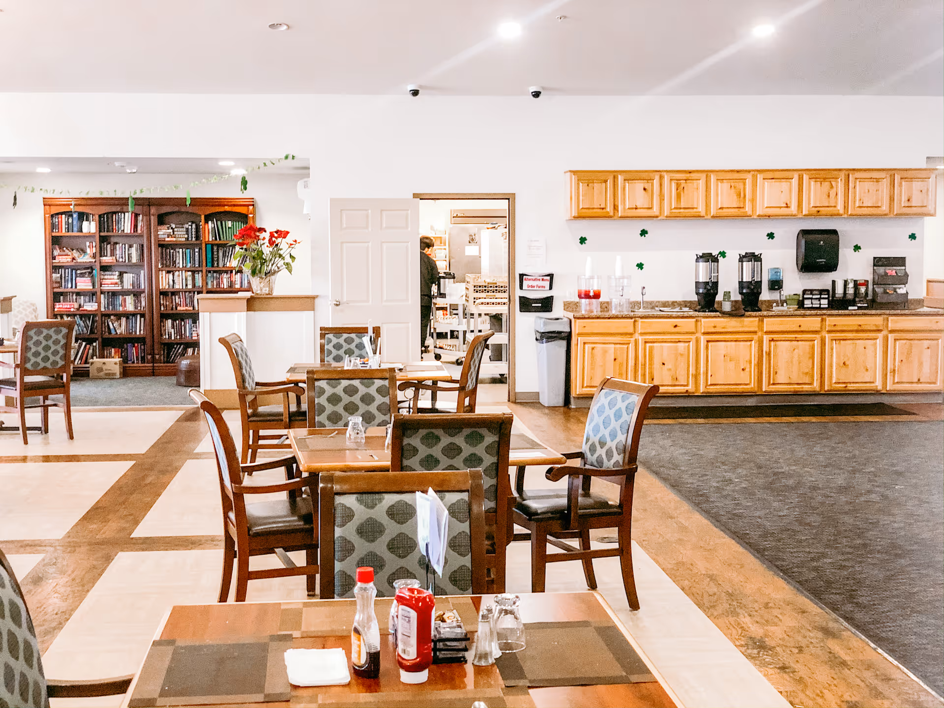 A bright communal dining room with wooden tables and upholstered chairs, condiments on a foreground table, a kitchenette with wood cabinets to the right and bookshelves in the back.