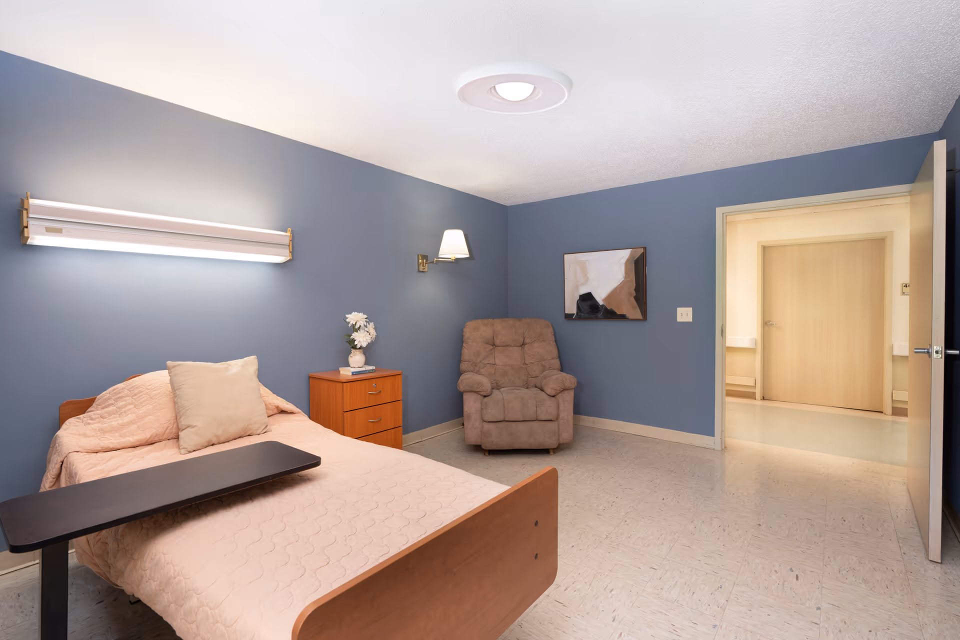 A tidy senior bedroom with a single bed, overbed table, nightstand with flowers, recliner chair, and an open doorway against blue walls.