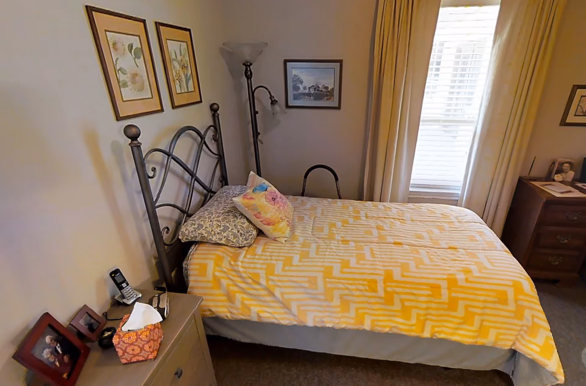A cozy bedroom with a single bed covered in a yellow and white patterned blanket and two pillows. The bed has a black metal headboard. There is a wooden nightstand with framed photos, a tissue box, and a cordless phone. The room has framed artwork on the walls, a floor lamp, and a window with white blinds and beige curtains.