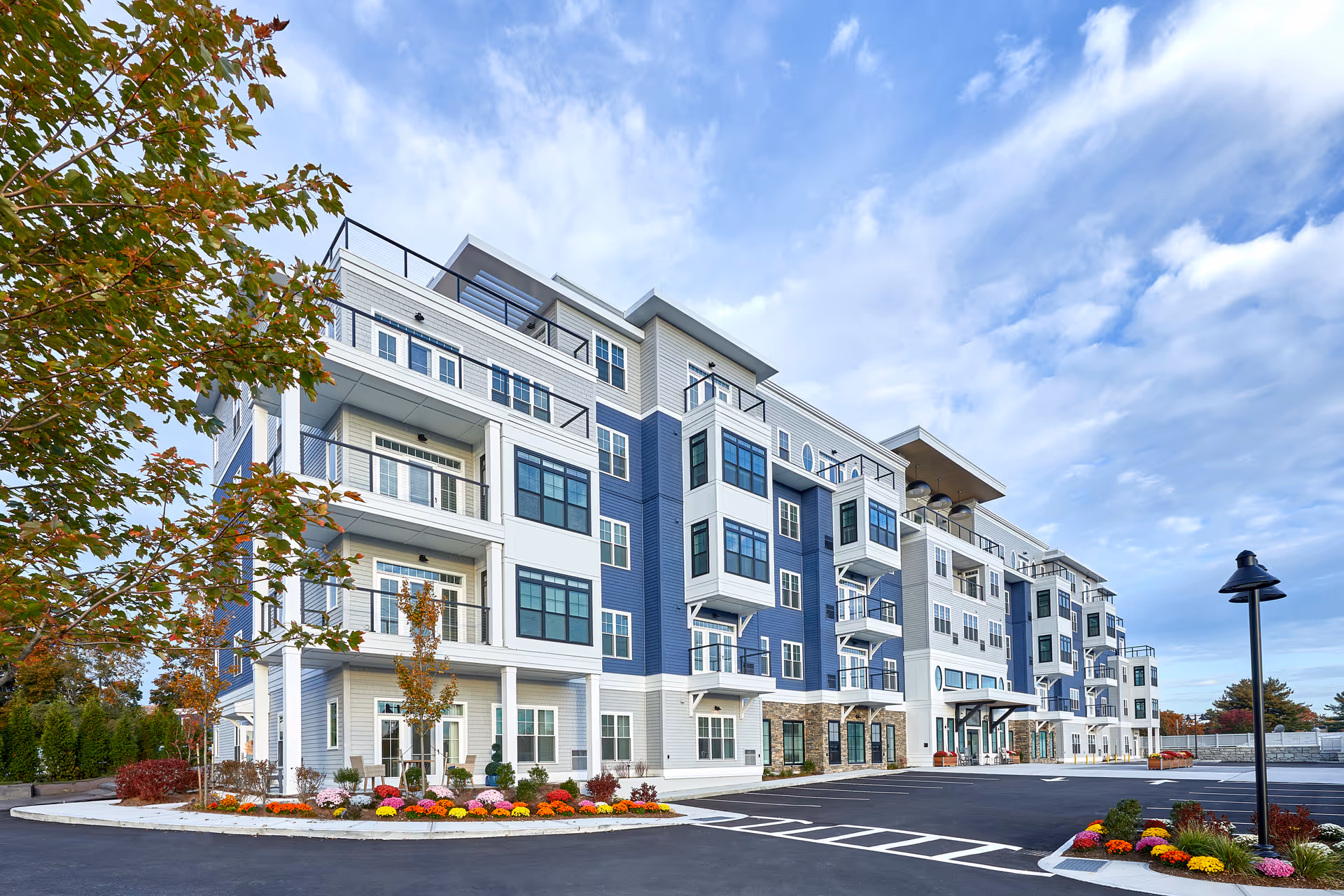 Front exterior of a multi-story blue-and-white senior living building with balconies, parking lot, and landscaped flower beds under a partly cloudy sky.