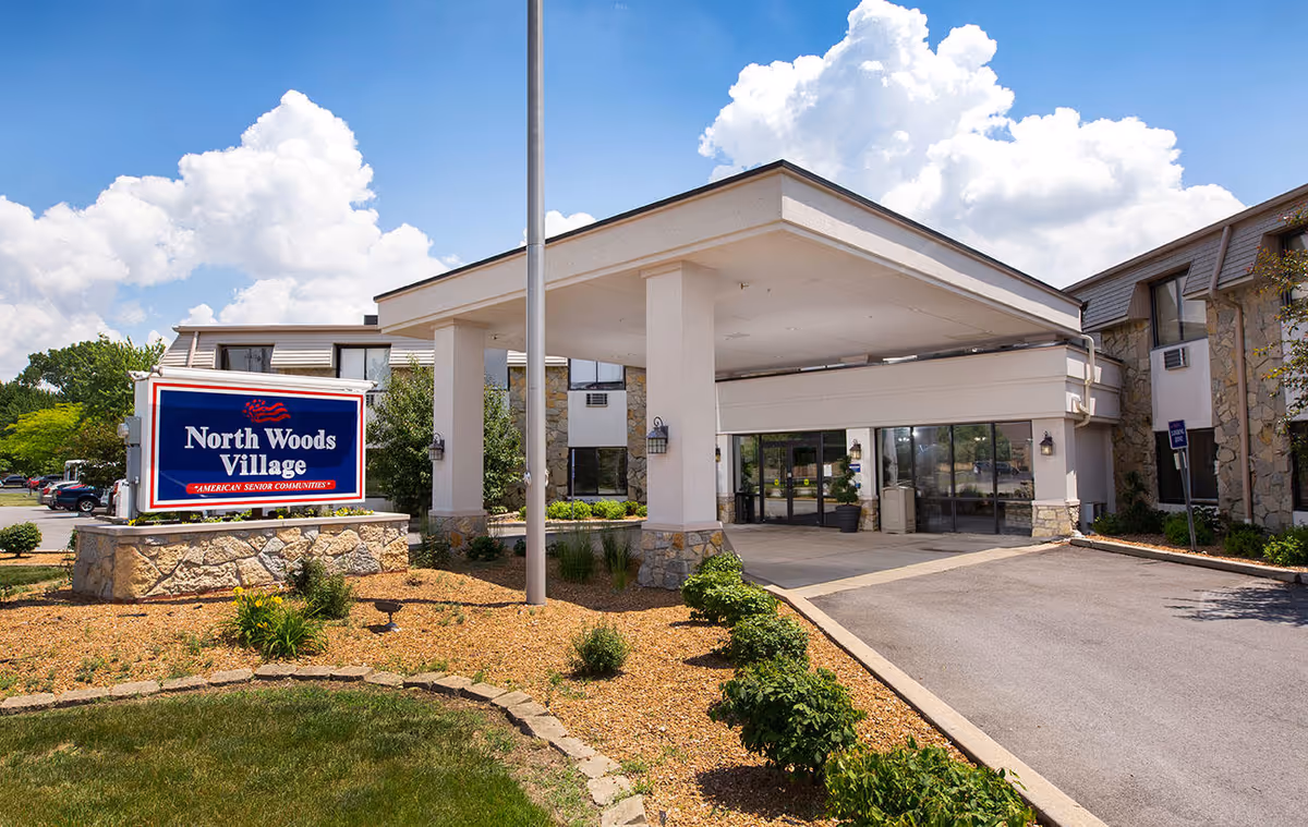 Exterior view of North Woods Village senior living facility entrance with a covered drop-off area, stone and stucco building facade, landscaped garden with bushes and mulch, and a clear blue sky with clouds.