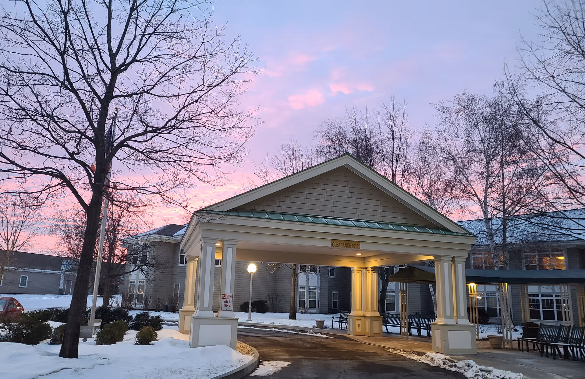 Entrance driveway of Massry Assisted Living Residence at dusk with a covered drop-off area supported by columns. Snow covers the ground and leafless trees surround the building. The sky is a mix of pink and purple hues.