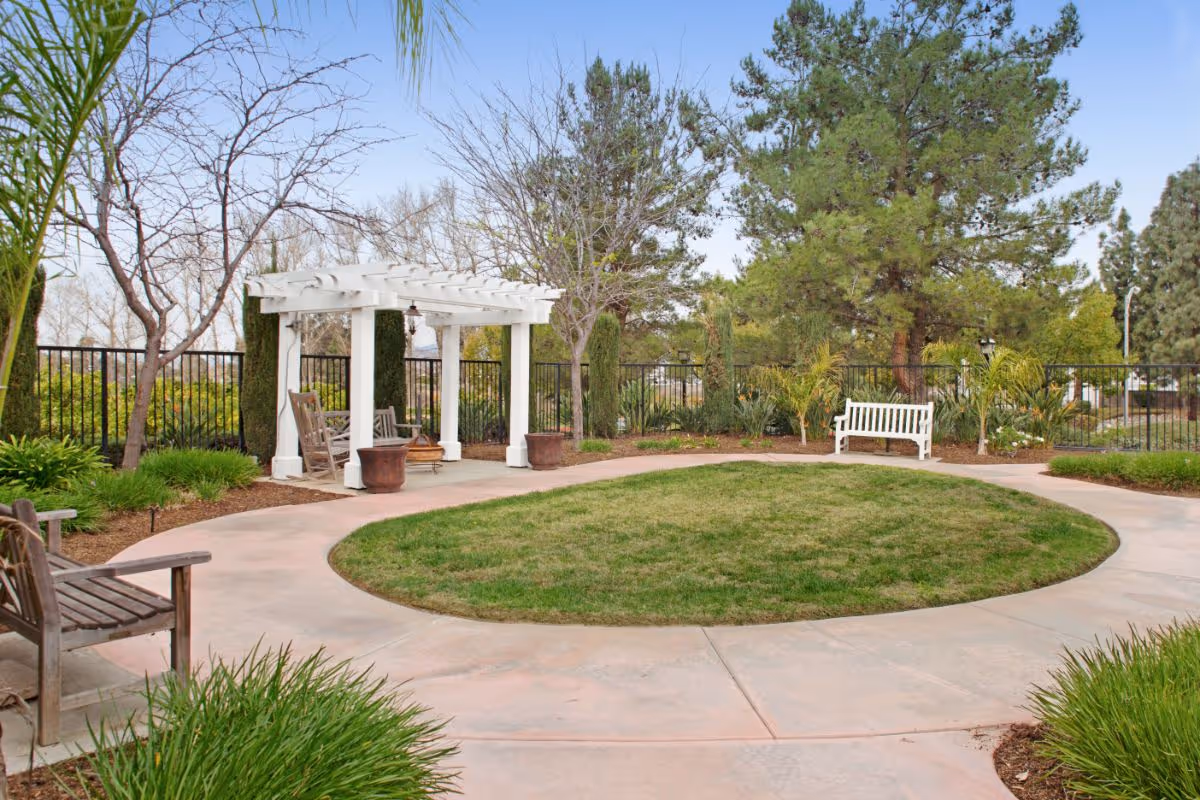 Circular paved courtyard with a grassy center, a white pergola, benches, and surrounding trees and landscaping.