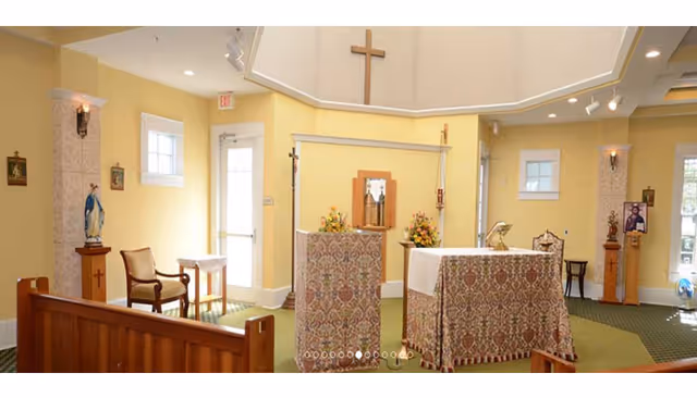 Interior view of a small chapel or prayer room with yellow walls, a wooden pew, religious statues, floral arrangements, a cross mounted on the wall, and an altar covered with a patterned cloth.