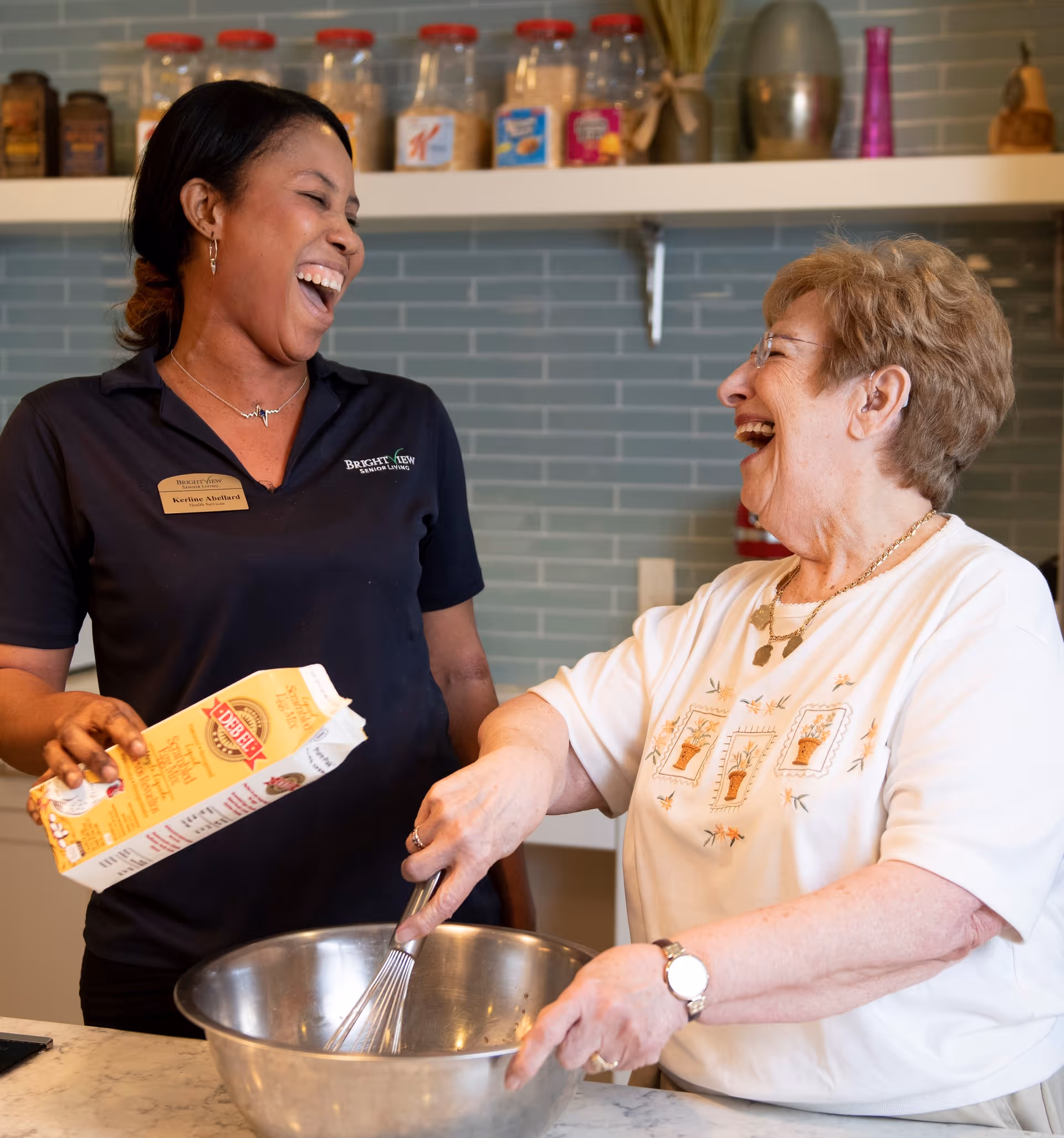 Two women laugh together in a kitchen as one stirs a large mixing bowl and the other pours ingredients.