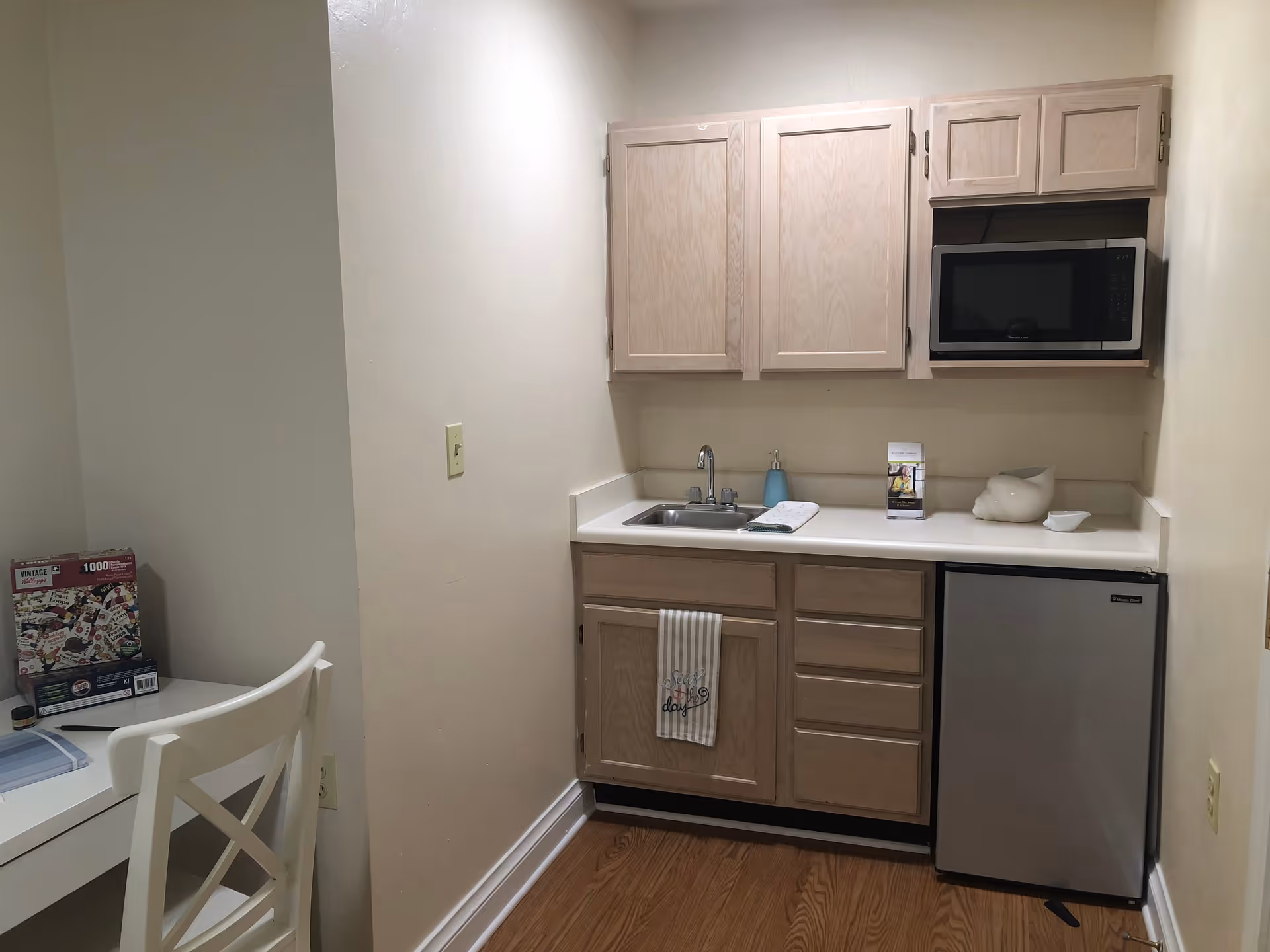Small kitchenette area with light wood cabinets, a stainless steel mini refrigerator, a microwave, a sink with a soap dispenser, and a towel hanging on the cabinet door. To the left, there is a white chair and a white table with a puzzle box and some small items on it. The floor is wooden and the walls are light-colored.
