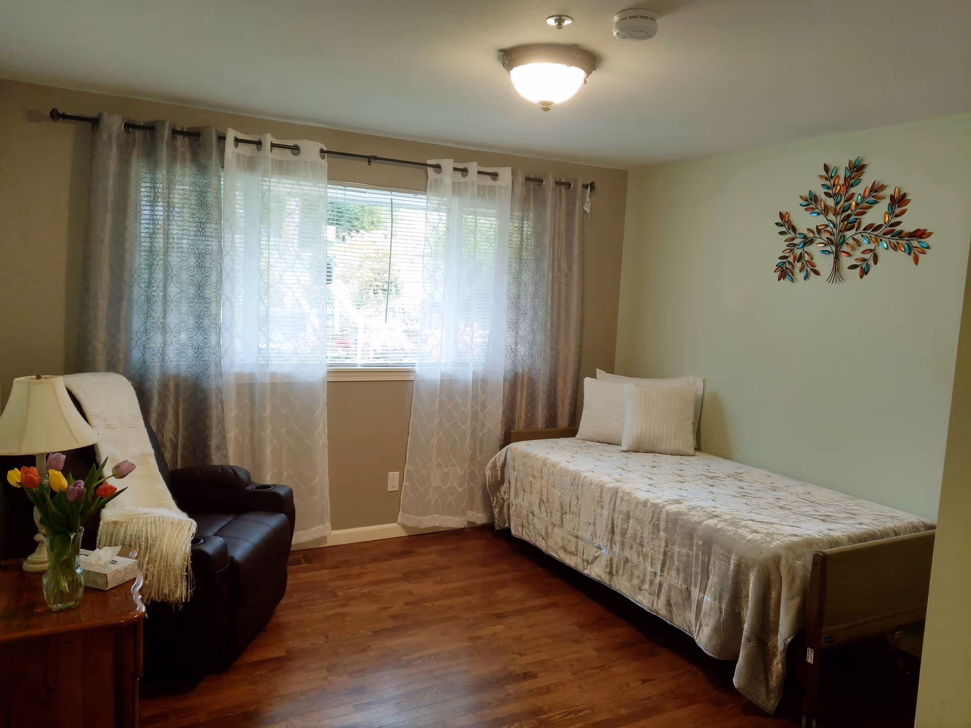 A cozy bedroom with a single bed covered in a light-colored bedspread and two pillows. There is a brown recliner chair with a white throw draped over it next to a wooden side table holding a lamp, a vase of colorful tulips, and a box of tissues. The room has wooden flooring, a window with sheer and patterned curtains, and a decorative metal tree wall art on a light green wall.