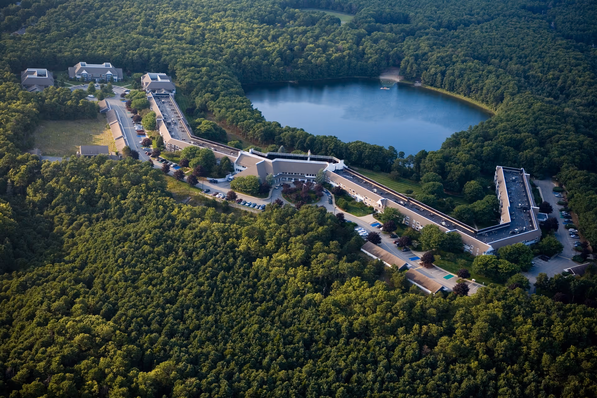 Aerial view of Thirwood Place senior living facility surrounded by dense forest with a large pond nearby. The building complex is long and angular with multiple parking areas and driveways.