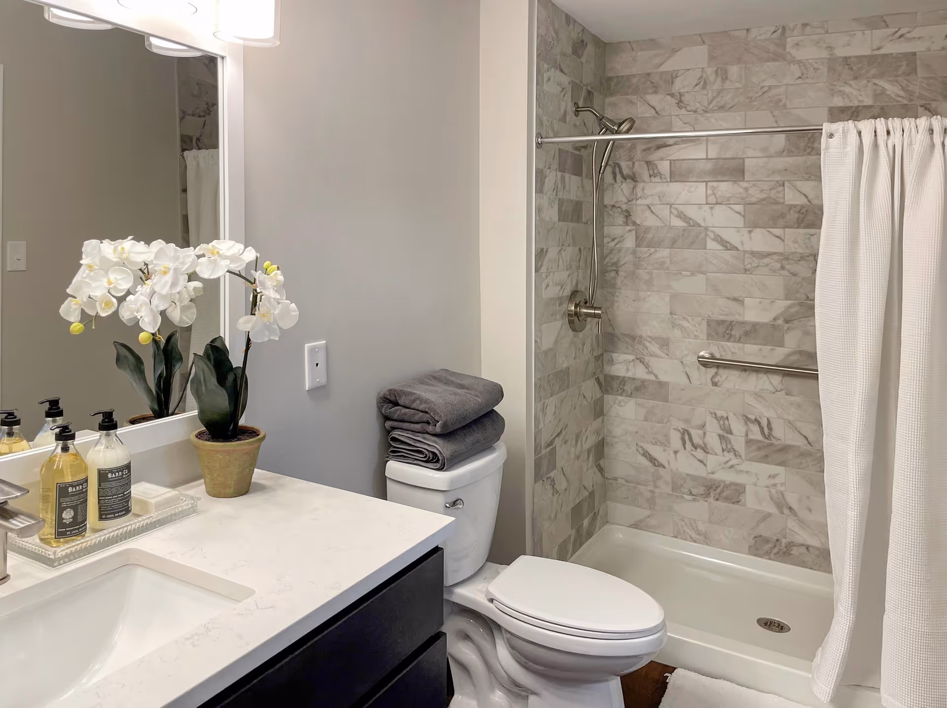 A clean and modern bathroom featuring a white countertop with a built-in sink, a potted white orchid, and soap dispensers. There is a toilet with two folded gray towels on top, and a shower area with a white curtain and marble-patterned tile walls. A large mirror is mounted above the sink.
