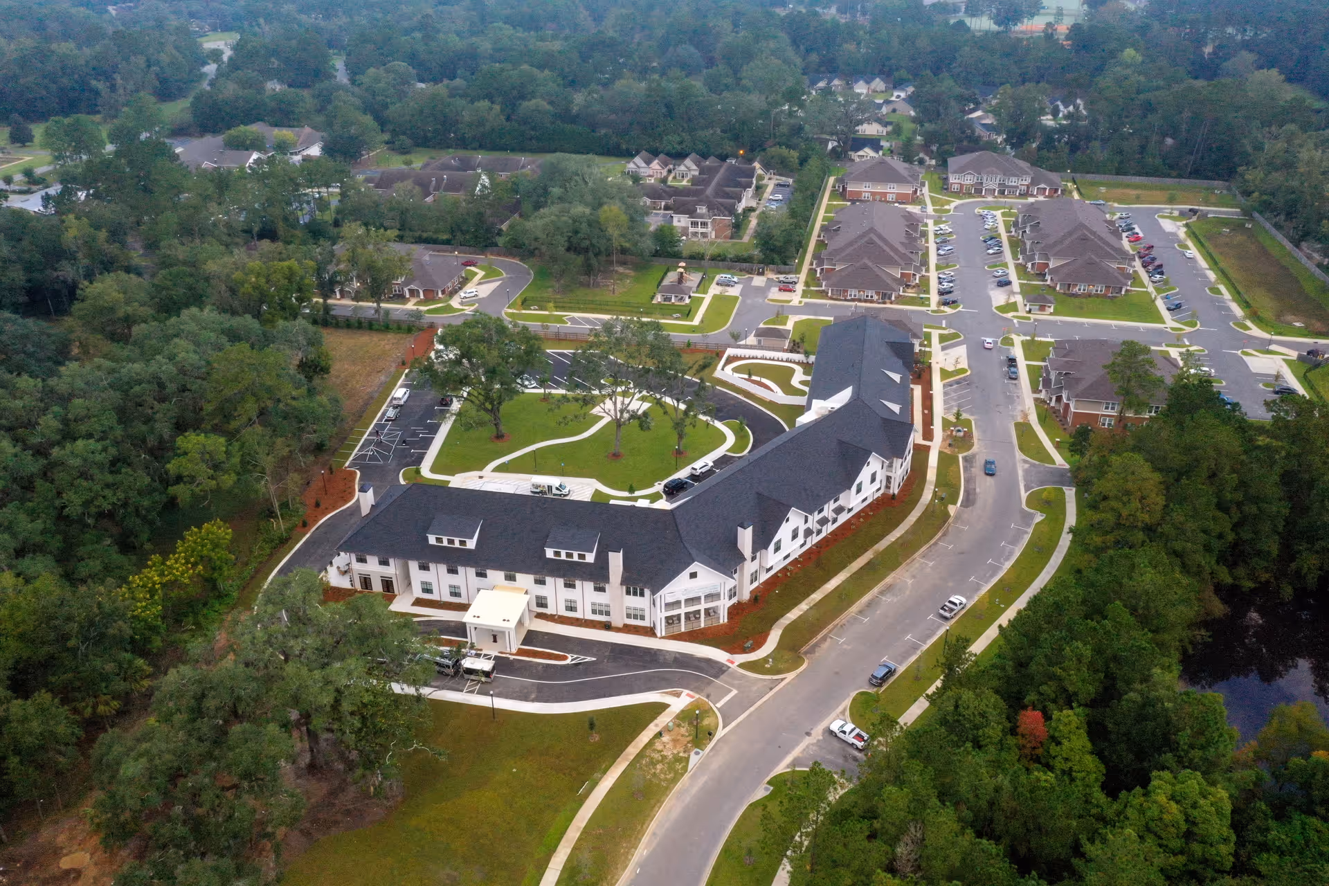 Aerial view of The Residence at Oak Grove Assisted Living & Memory Care facility showing multiple buildings with dark roofs, parking lots, roads, and surrounding greenery including trees and a small pond.