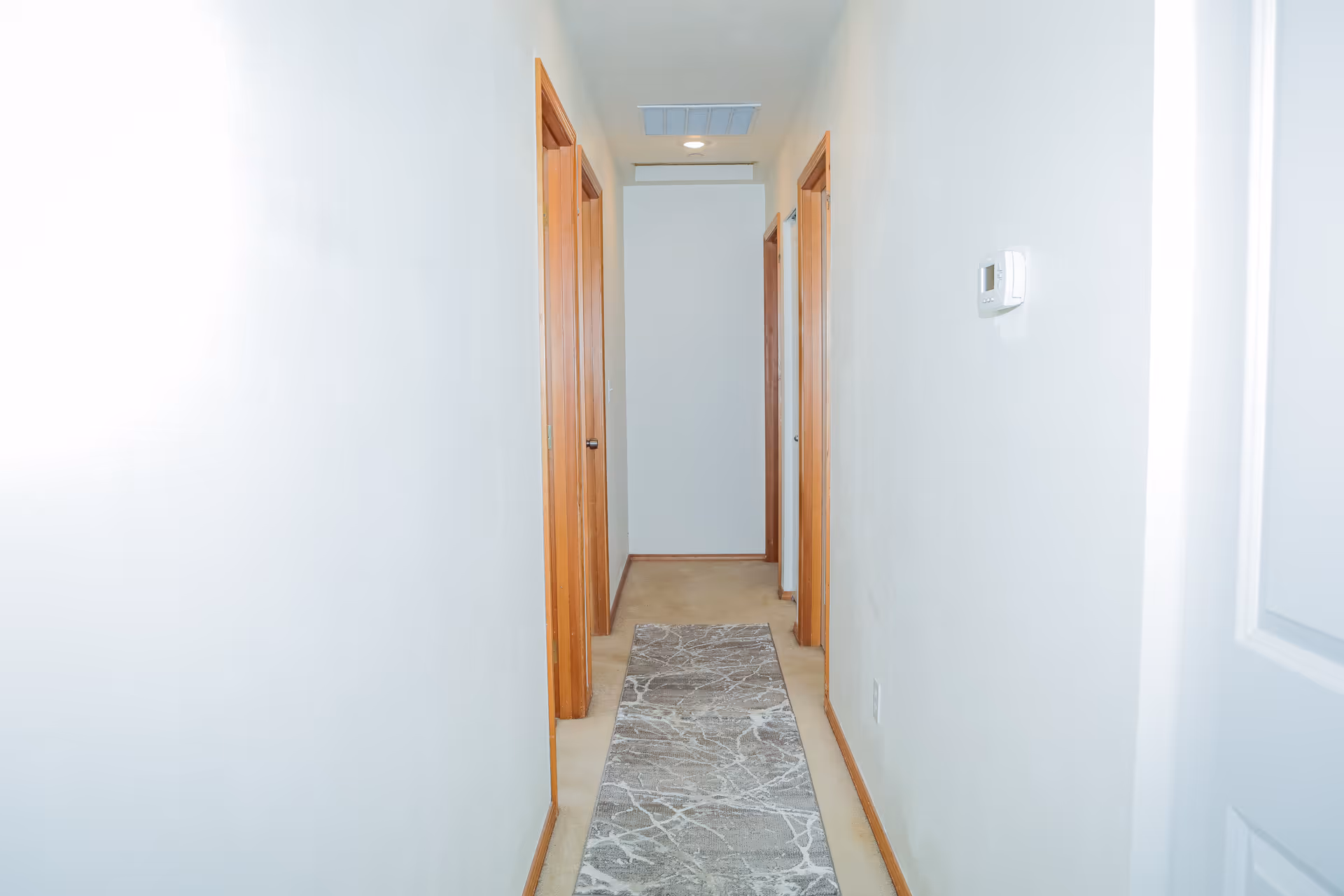 A narrow hallway with white walls and light brown wooden door frames on both sides. A gray patterned runner rug is placed on the beige carpet floor. A thermostat is mounted on the right wall, and a ceiling light fixture is visible at the end of the hallway.