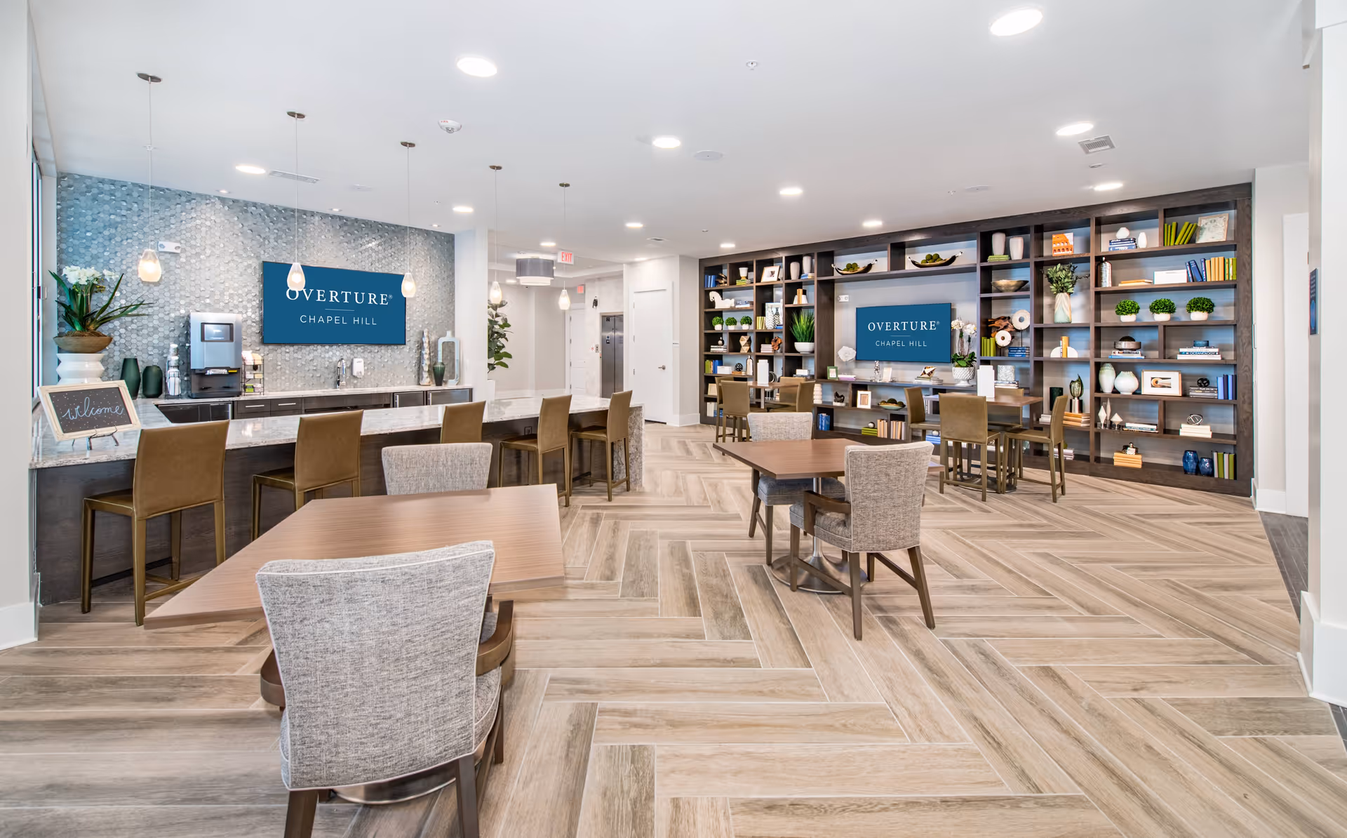 A modern, well-lit common area in Overture Chapel Hill featuring a bar counter with high chairs, several tables with upholstered chairs, and a large built-in bookshelf decorated with plants, books, and decorative items. Two screens display the Overture Chapel Hill logo on the back wall.