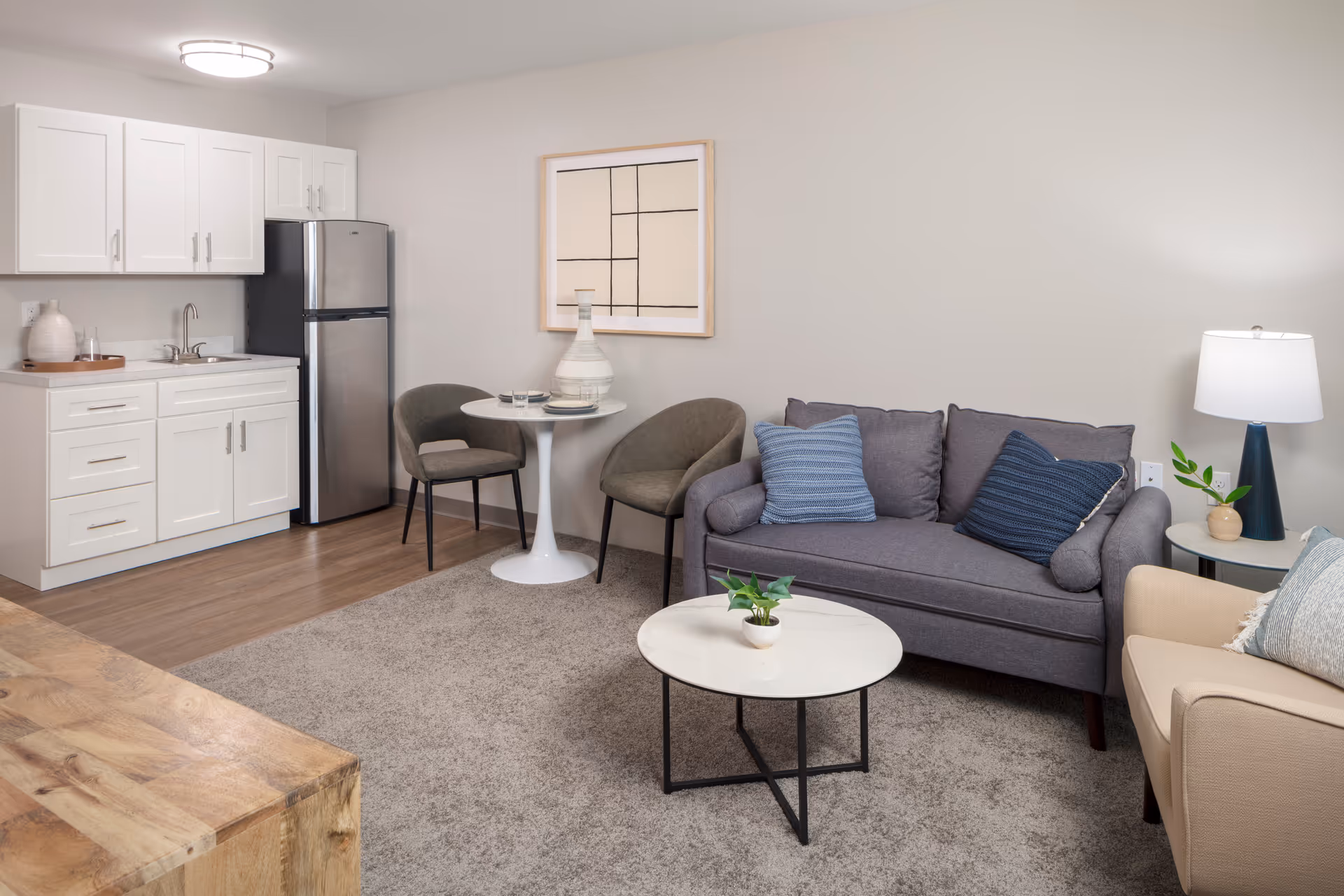 A cozy living area with a small kitchen in the background. The kitchen has white cabinets, a stainless steel refrigerator, and a sink. In front of the kitchen is a small round white table with two gray chairs. The living area features a gray sofa with blue pillows, a beige armchair with a light blue pillow, a round white coffee table with a small plant, and a side table with a lamp and another small plant. The floor is a combination of wood and carpet, and a framed abstract artwork hangs on the wall.