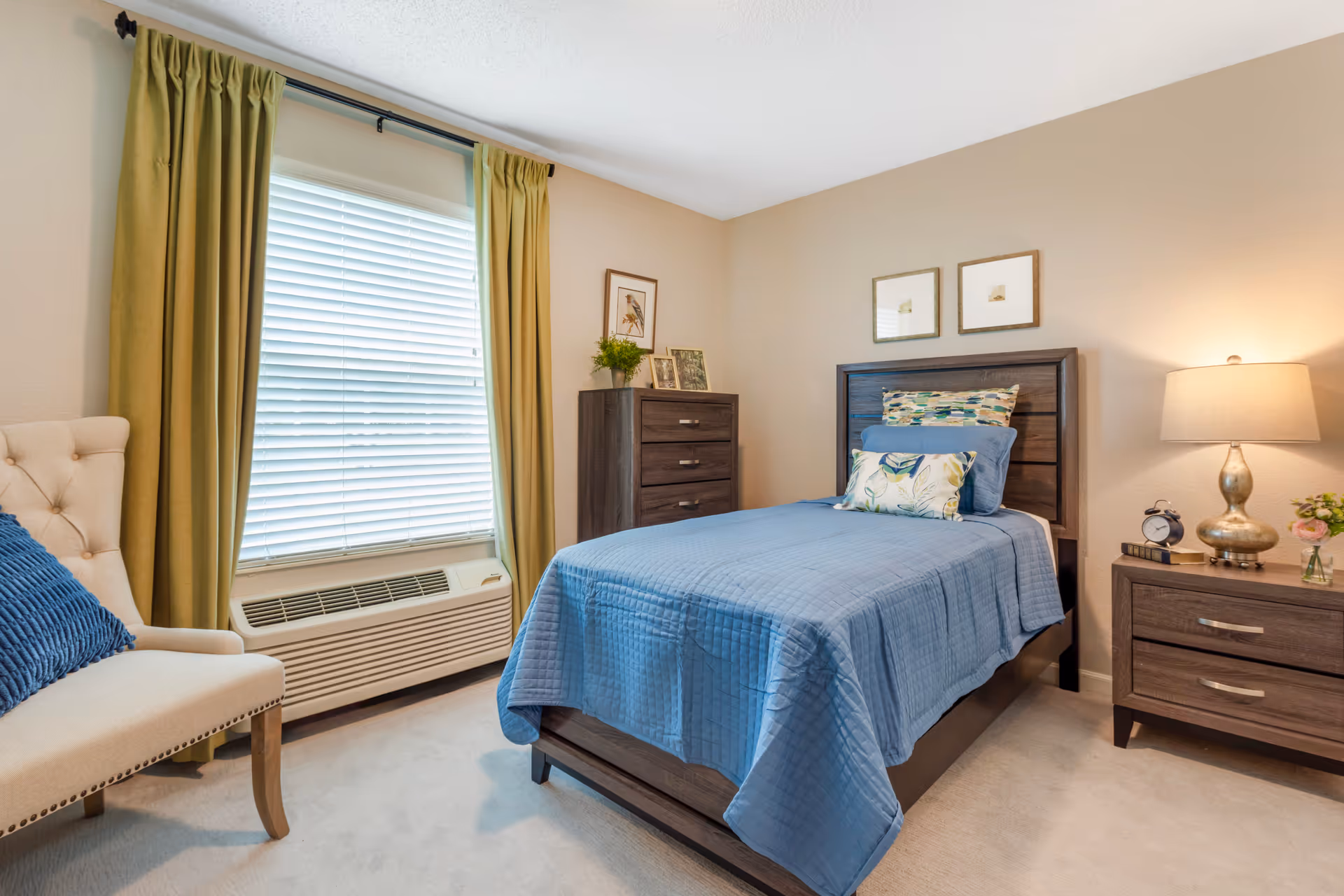 A neatly staged bedroom with a twin bed dressed in blue linens, a nightstand with lamp and alarm clock, dresser, window with green curtains, and an upholstered chair.