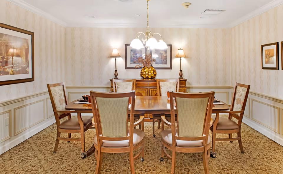 Well-lit dining room with a wooden table surrounded by six chairs, table settings, lamps, and framed artwork.