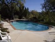 Outdoor swimming pool surrounded by concrete deck with white plastic chairs and trees in the background under a clear blue sky.