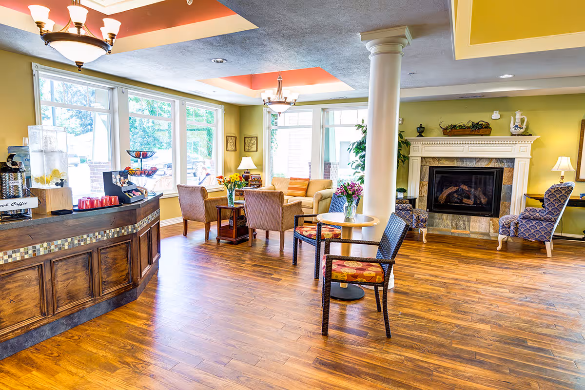 A bright and welcoming common area in a senior living facility with wooden floors, a fireplace, comfortable armchairs, a sofa, small tables with flower vases, large windows letting in natural light, and a coffee station on the left side.