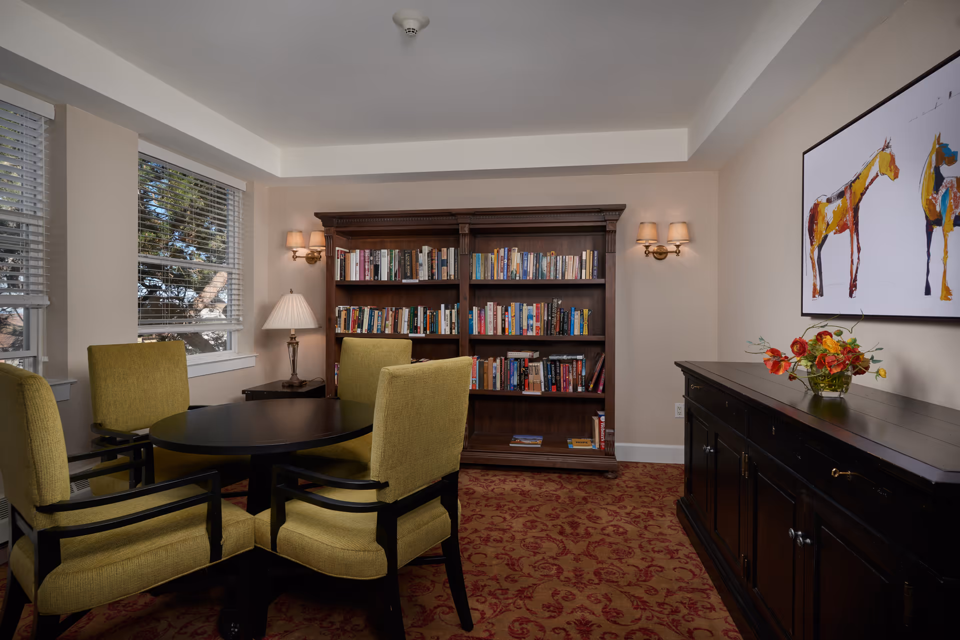 Small common room with a round table surrounded by four green chairs, a large bookshelf, sideboard with flowers, and wall art.