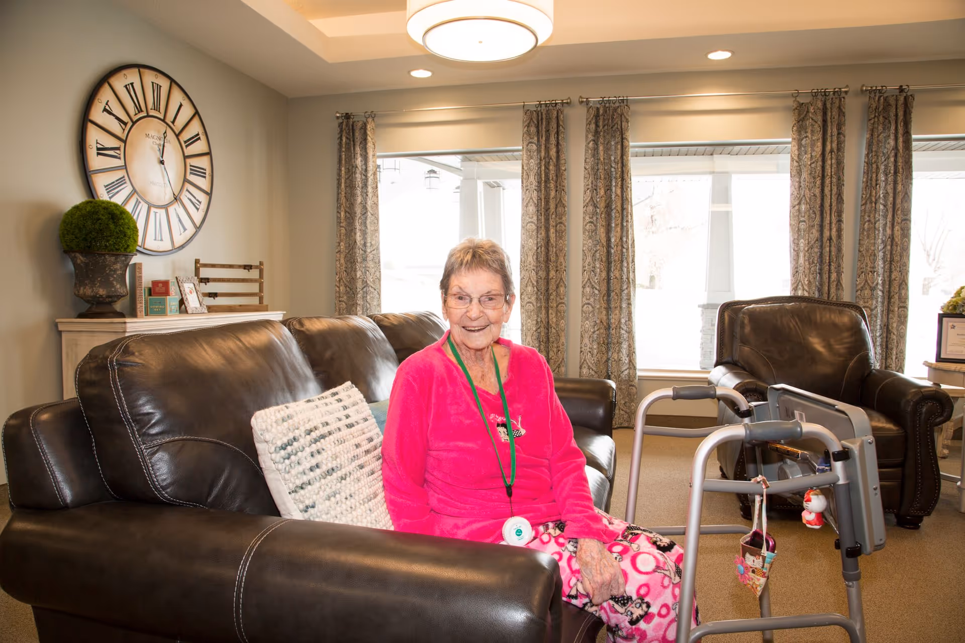 An older woman wearing pink sits on a leather sofa in a well-lit living room with a walker beside her.