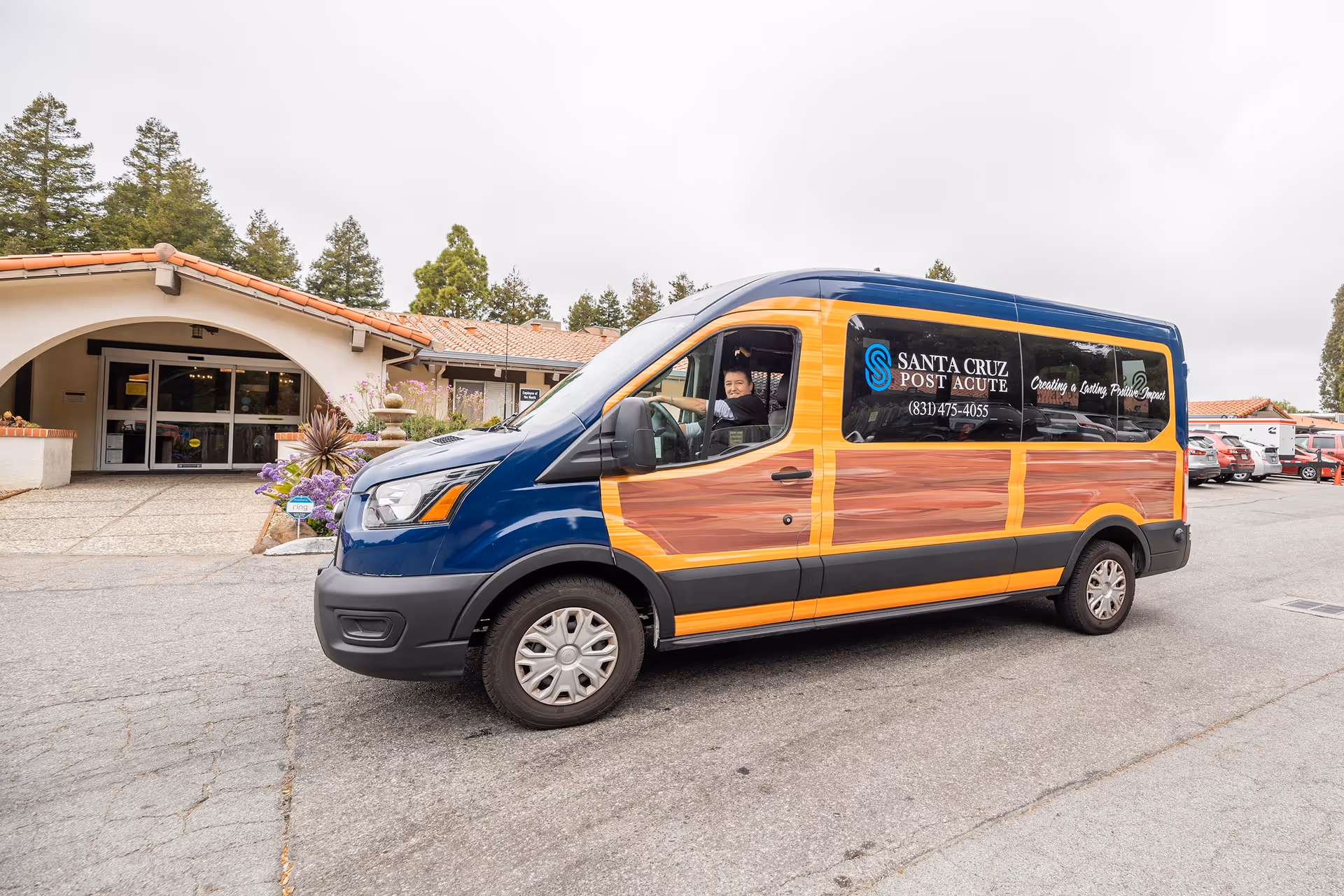 A blue and orange shuttle van with wood panel design parked in front of a building with a tiled roof and arched entrance. A person is sitting in the driver's seat smiling. The van has the text 'Santa Cruz Post Acute' and a phone number on the side window.