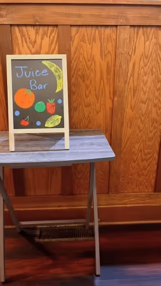 A small chalkboard sign on a wooden table with a wood-paneled wall in the background. The sign reads 'Juice Bar' and is decorated with colorful drawings of various fruits including a banana, orange, strawberry, cherry, and lemon.